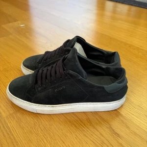 Svarta sneakers från Axel Arigato - Säljer ett par stilrena svarta sneakers från Axel Arigato i mocka med vita platta sulor och svarta skosnören. Klassisk låg modell med rund tå och diskret logga på sidan. Perfekta för en clean och modern look. Påsen medföljer