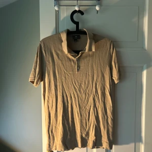Beige pikétröja från H&M - Säljer en beige pikétröja från H&M i slim fit-modell. Tröjan har korta ärmar, klassisk krage och knappar framtill. Perfekt för en stilren och enkel look. Materialet känns mjukt och bekvämt.