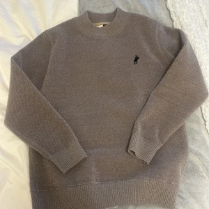 Grå stickad tröja från Ralph Lauren - Säljer en klassisk grå stickad tröja från Ralph Lauren med rund halsringning och broderad logga på bröstet. Perfekt för lager-på-lager och passar till det mesta. Mjuk och skön kvalitet.