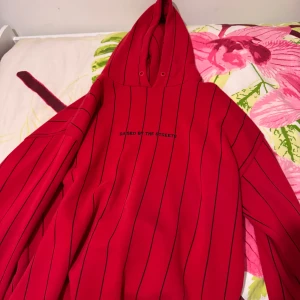 Röd hoodie med svarta ränder från black squad  - Säljer en röd hoodie med svarta tunna ränder och texten 'RAISED BY THE STREETS' på bröstet. Tröjan har huva och långa ärmar, perfekt för en avslappnad stil. Baggy stil