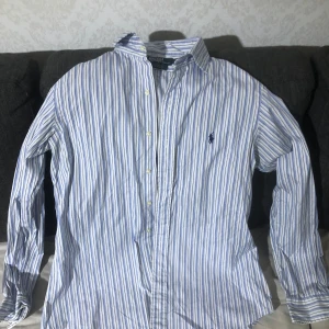 Blårandig skjorta från Ralph Lauren - Klassisk skjorta med blå och vita ränder från Ralph Lauren. Skjortan har lång ärm, knappar framtill och den ikoniska broderade loggan på bröstet. Perfekt för en stilren och tidlös look.