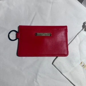 Röd korthållare  Emporio Armani - Säljer en stilren röd korthållare från Emporio Armani i skinn. Den har ett metallmärke framtill, nyckelring och ett kuvertformat fack med tryckknapp. Perfekt för kort och småsaker, smidig att ha med sig överallt.