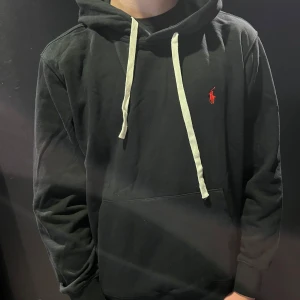 Svart hoodie från Polo Ralph Lauren - Svart hoodie med vit dragsko och röd broderad Polo-logga på bröstet. Klassisk känguruficka framtill och mjuk bomullskänsla. Perfekt för en avslappnad och stilren look.