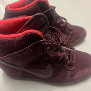 Vinröda Nike sneakers med högt skaft - Säljer ett par vinröda Nike sneakers med högt skaft och matchande röda detaljer. Skorna har snörning och kommer med extra skosnören. Perfekta för dig som vill sticka ut med en unik färg på dina sneakers.