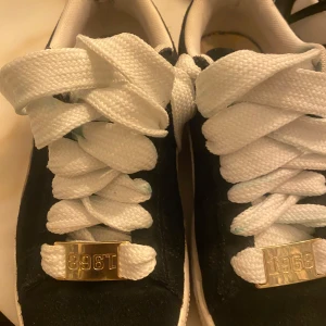 Svarta Puma Suede sneakers med vita detaljer - Säljer ett par klassiska Puma Suede sneakers i svart mocka med vita detaljer och breda vita skosnören. Guldiga metalldetaljer framtill och Puma-logga i guld på sidan. Perfekta för dig som gillar retrostil och streetwear.