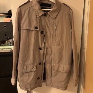 Beige jacka/trenchcoat från Zara Man med bälte - Säljer en beige jacka/trenchcoat från Zara Man med klassisk krage, knappar framtill, dragkedjeficka på bröstet och bälte i midjan. Jackan har långa ärmar och flera praktiska fickor. Perfekt för vår och höst. Liten fläck på framsidan men inte jätte synlig.