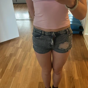 Grå slitna jeansshorts - Säljer ett par grå jeansshorts med låg midja och snörning fram. Shortsen har en sliten detalj på ena låret och klassiska fickor både fram och bak. Perfekta för sommaren! Orginal pris 200kr 