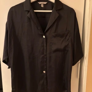 Svart kortärmad satin-skjorta från H&M - Säljer en svart satin-skjorta från H&M i storlek S. Skjortan har korta ärmar, klassisk krage, knäppning framtill och en bröstficka. Tillverkad i 100% polyester vilket ger en lätt och mjuk känsla.