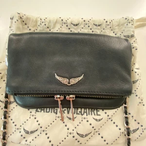 Svart axelväska från Zadig & Voltaire - Snygg svart axelväska i skinn från Zadig & Voltaire med silverfärgade detaljer och kedjerem. Väskan har dubbla dragkedjor framtill, ett dekorativt vingemblem och en extra ficka med dragkedja på baksidan. Den är lite rostig på kedjorna därmed priset ❤️🥰