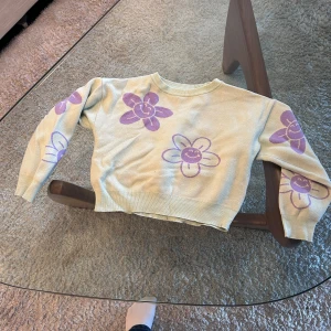 Limegrön stickad tröja med lila blommor från WOOW - Superfin  stickad tröja från WOW (Cubus) med stora lila blommor och glada ansikten. Tröjan har en croppad passform och långa ärmar. Perfekt för dig som vill sticka ut lite extra!