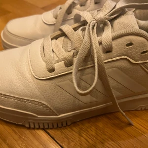 Vita sneakers från Adidas - Säljer ett par fräscha vita sneakers från Adidas med klassiska ränder på sidan. Skorna har snörning och är tillverkade i syntetmaterial med en platt sula. Perfekta för en clean och sportig look.