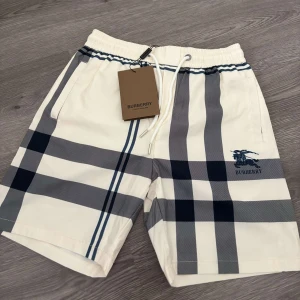 Vita rutig shorts från Burberry - Snygga vita shorts från Burberry med klassiskt rutigt mönster i grått och blått. Shortsen har snörning i midjan och sidofickor. Perfekta för sommaren och har en broderad Burberry-logga på ena benet. Topp kvalite, hör av er vid frågor.:)