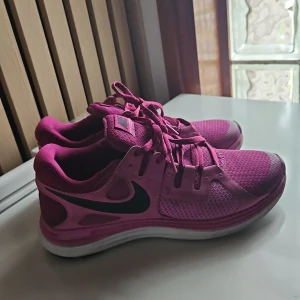 Rosa Nike sneakers med svart swoosh - Säljer ett par snygga rosa Nike sneakers med svart swoosh och vita sulor. Skorna har meshpartier för bra ventilation och klassisk snörning. Perfekta för träning eller vardagsstil.