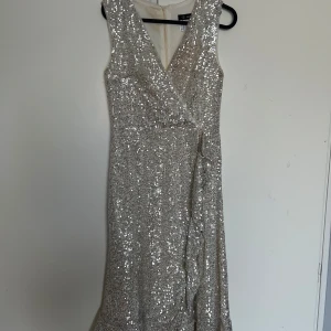 Silver glittrig långklänning med omlott - Säljer en snygg silverfärgad långklänning med massor av paljetter. Klänningen är ärmlös, har v-ringning och omlott-detalj framtill. Perfekt för fest eller nyår! Supersnygg och glittrar mycket.