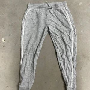 Grå mjukisbyxor med vita ränder från Gina Tricot - Säljer ett par grå mjukisbyxor från Gina Tricot med vita ränder längs sidorna. Byxorna har resår i midjan med snörning och är tillverkade i mjuk bomull. Perfekta för en avslappnad stil.