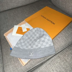 Grå rutig mössa från Louis Vuitton - Snygg grå mössa från Louis Vuitton med klassiskt rutigt mönster och diskret LV-logga framtill. Tillverkad i mjukt material som känns lyxigt och bekvämt. Perfekt accessoar för att lyfta din stil.