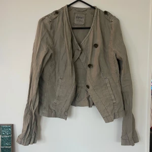 Beige kavajjacka från Esprit - Säljer en beige kavajjacka från Esprit med asymmetrisk knäppning och dekorativa axelremmar. Jackan har lång ärm, två fickor fram och en lätt rynkad detalj baktill. Tillverkad i ett tunt, linne material. Perfekt för vår och höst.