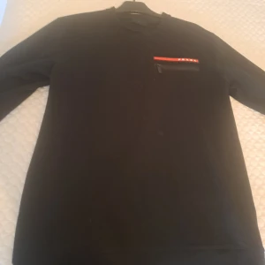 Prada tröja - Svart långärmad tröja med en liten ficka på bröstet som har dragkedja och en röd detalj med stjärnor. Enkel och stilren modell som passar till mycket. Storlek L