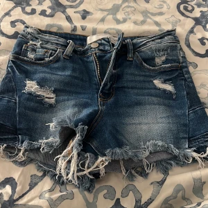 Blå jeansshorts med fransar från Bridge by Gly - Säljer ett par blå jeansshorts från Bridge by Gly med slitna detaljer och fransig kant nertill. Shortsen har fem fickor och är högmidjade. Materialet är en mix av bomull, polyester och spandex för lite stretch.