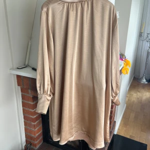 Champagnefärgad skjortklänning i satin - Säljer en champagnefärgad skjortklänning i glansigt satinmaterial. Klänningen har lång ärm med lätt puff och manschett, klassisk krage och rak passform. Perfekt för dig som gillar stilrena och eleganta plagg.