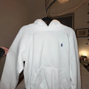 Vit hoodie från Polo Ralph Lauren - Säljer en klassisk vit hoodie från Polo Ralph Lauren med broderad blå logga på bröstet. Tröjan har huva och en stor magficka framtill. Perfekt för en clean och stilren look.