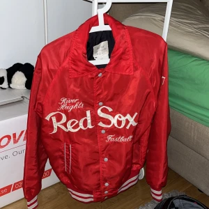 Röd baseballjacka Red Sox från Mid-West Sportswear - Riktigt snygg röd baseballjacka med Red Sox-brodyr framtill och vita detaljer. Jackan har knappar, ribbade muddar och krage samt klassisk sportig look. Perfekt för dig som gillar retrostil och sportmode.