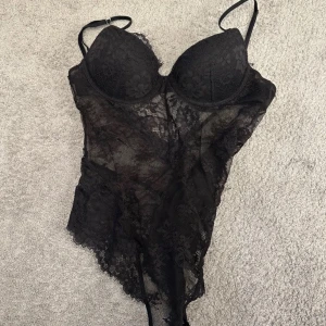 Svart spetsig bodysuit storlek 75A - Säljer en svart bodysuit i spets med tunna axelband och v-ringning. Bodyn har genomskinliga partier och en figurnära passform, perfekt att styla med jeans eller kjol. Snygg och trendig detalj till din garderob!