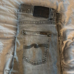 Grå jeans från Nudie Jeans - Säljer ett par grå jeans från Nudie Jeans med klassisk femficksdesign och svarta broderade vågor på bakfickorna. Jeansen har en rak passform och svart patch bak i midjan. Perfekta för en stilren vardagslook.  Modell: Lean Dean Strolek: W29 L34