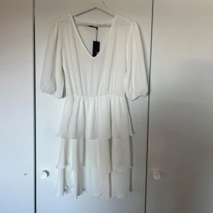 Vit volangklänning med v-ringning - Superfin vit klänning med v-ringning och trekvartsärmar. Klänningen har flera lager volanger i kjolen som ger en flowy känsla. Perfekt för sommarens alla tillfällen och passar även bra till studenten! Den är i bra skick då den inte är använd och prislappen är fortfarande kvar!✨🫧🩷