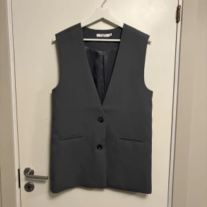Oversized grå väst från NA-KD - Stilren oversized grå väst från NA-KD med två knappar framtill och två fickor. Västen har en rak passform och är ärmlös, perfekt att styla över en skjorta eller t-shirt för en trendig look.