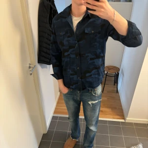 Blå camouflagemönstrad skjorta - Snygg blå camouflagemönstrad skjorta med långa ärmar och knappar framtill. Perfekt för en avslappnad stil. Skjortan har två bröstfickor med knappstängning. Brodering på bröstet och axeln. Storlek L men sitter mer som M. Jag är 185cm