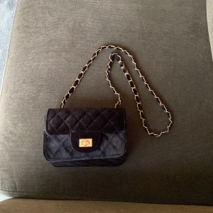 Svart quiltad axelväska med guldkedja - Snygg svart axelväska med quiltat mönster och guldfärgad kedja. Väskan har ett klassiskt lock med vridspänne i guld och är perfekt för att bära det viktigaste. Liten och smidig modell som passar till många tillfällen.