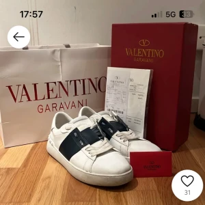 Valentino Garavani vita sneakers med svart band - Säljer ett par vita Valentino Garavani sneakers med ett brett mörkblått band över vristen och nitar på hälen. Skorna har rund tå, platt sula och snörning. Klassisk och stilren design med märkets logga på plösen. Bilderna är mina, bara tagna från en gammal annons. Priset gör att diskutera.