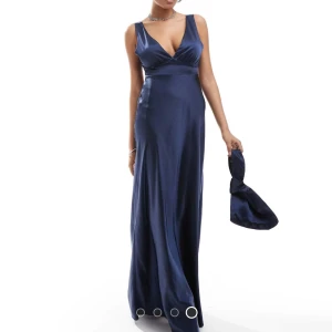 Marinblå långklänning med rosett från Six Stories - Elegant marinblå långklänning med breda axelband och stor rosett i ryggen. Klänningen har maxilängd och dekorativa ringdetaljer längst ner. Perfekt för bal eller festliga tillfällen.