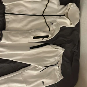 Vit hoodie och byxor från Nike - Säljer ett set med vit hoodie och matchande byxor från Nike. Hoodien har dragkedja, huva och svarta detaljer samt en ficka på ärmen. Byxorna har resår i midjan och svarta snören. Perfekt för en sportig och avslappnad stil.