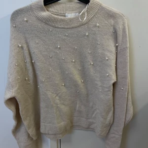 Beige stickad tröja med pärlor - Superfin beige stickad tröja med pärldetaljer på framsidan. Tröjan har rund halsringning, långa ärmar och ribbade muddar. Perfekt för att addera lite extra till din outfit!