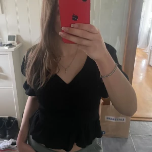 Svart omlottopp med volang - Säljer en svart omlottopp med v-ringning och volang nertill. Toppen har korta puffärmar och en feminin passform som framhäver midjan. Endast använd en gång, jättebra skick!💕Storlek XS men är lite större, så skulle säkert sitta som S eller M!