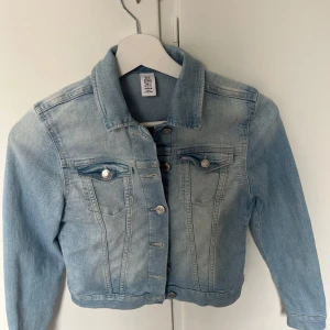 Ljusblå jeansjacka från H&M - Säljer en klassisk ljusblå jeansjacka från H&M  med silverfärgade knappar och två bröstfickor med ficka. Jackan har krage och långa ärmar, perfekt för vår och sommar. Snygg och enkel att matcha till olika outfits.