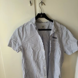 Randig kortärmad skjorta från Marwin - Säljer en ljusblå och vit randig kortärmad skjorta från Marwin. Skjortan har klassisk krage, knappar framtill och diskret broderad text på bröstet. Perfekt för en stilren och avslappnad look.