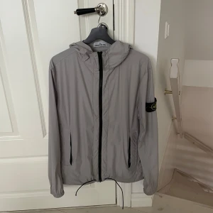 Ljusgrå vindjacka från Stone Island - Snygg ljusgrå vindjacka från Stone Island med huva och svarta detaljer. Jackan har dragkedja framtill, två sidofickor med dragkedja och klassisk logotyp-patch på ärmen. Perfekt för vår, höst och en skön sommar kväll.