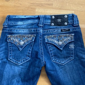 Blå low waist, bootcut Miss Me jeans med broderade bakfickor - Säljer ett par blå jeans från Miss Me med snygga broderade detaljer och pärlor på bakfickorna. Jeansen är mycket low waist och bootcut. Älskar dessa jeans då de är skit snygga men säljer på grund av att de är lite för små. Jag är 168cm och de är perfekta i längden. Jag köpte de second hand i london förra året och har bara använt dem några få gånger.