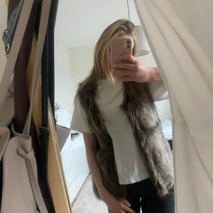 Gråbrun pälsväst - Säljer en snygg gråbrun pälsväst med naturlig look. Västen är ärmlös och har en rak passform som passar perfekt över en t-shirt eller tröja. Fejkpäls!! Perfekt för att addera lite extra stil till din outfit.