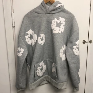 Grå hoodie med vita blommönster från Denim Tears - Säljer en grå hoodie från Denim Tears med unika vita blommönster över hela tröjan. Hoodien har en klassisk känguruficka och ribbade muddar vid ärmslut och nederkant. Perfekt för dig som vill sticka ut med en cool streetwear-look.