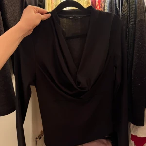 Svart långärmad omlottopp - Snygg svart topp med lång ärm och omlottdesign framtill. Djup v-ringning och elegant drapering vid halsen. Perfekt för en stilren look.