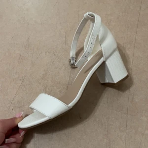 Vita sandaler med klack - Säljer ett par vita sandaler från XIT med bred rem över tårna och justerbart ankelband med spänne. Skorna har en stabil blockklack och öppen tå. Används ett par gånger men har inga skador, storlek 36. 