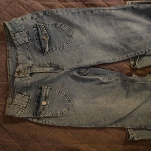 Grå jeans med stora fickor och knappar - Säljer ett par grå jeans med unika stora fickor framtill och dekorativa knappar. Byxorna har rak passform och klassisk jeanslook, perfekta för dig som vill sticka ut lite extra. Materialet är slitstarkt denim och färgen är ljusgrå. Det är storlek XL men skulle säga att den är mer som L 