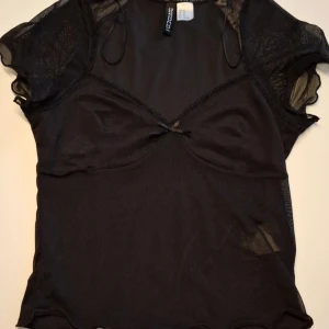 Svart mesh topp från H&M Divided - Säljer en svart mesh topp från H&M Divided i storlek XS. Toppen har korta ärmar i transparent mesh, liten rosett framtill och dekorativa spetsdetaljer.