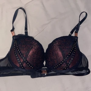 Svart och vinröd bh med spets och silvriga prickar - Säljer en snygg bh i svart och vinrött med spetsdetaljer och dekorativa nitar. Bh:n har justerbara axelband och stängning bak. Perfekt för dig som vill ha något extra fint under kläderna.