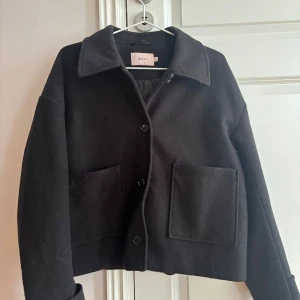 Svart kappa från Nelly - Stilren svart kappa från Nelly med klassisk krage och stora framfickor. Jackan har knäppning med knappar framtill, perfekt för dig som gillar en enkel och tidlös look!