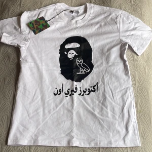 Vit t-shirt från Bape x OVO - Vit t-shirt från Bape i samarbete med OVO. Stor svart print med Bape-apa och OVO-uggla på bröstet samt text på arabiska. Klassisk rund hals och korta ärmar. Ikonisk camo-tag och detaljerad etikett. Helt fabriksny och aldrig provad. Storlek L.